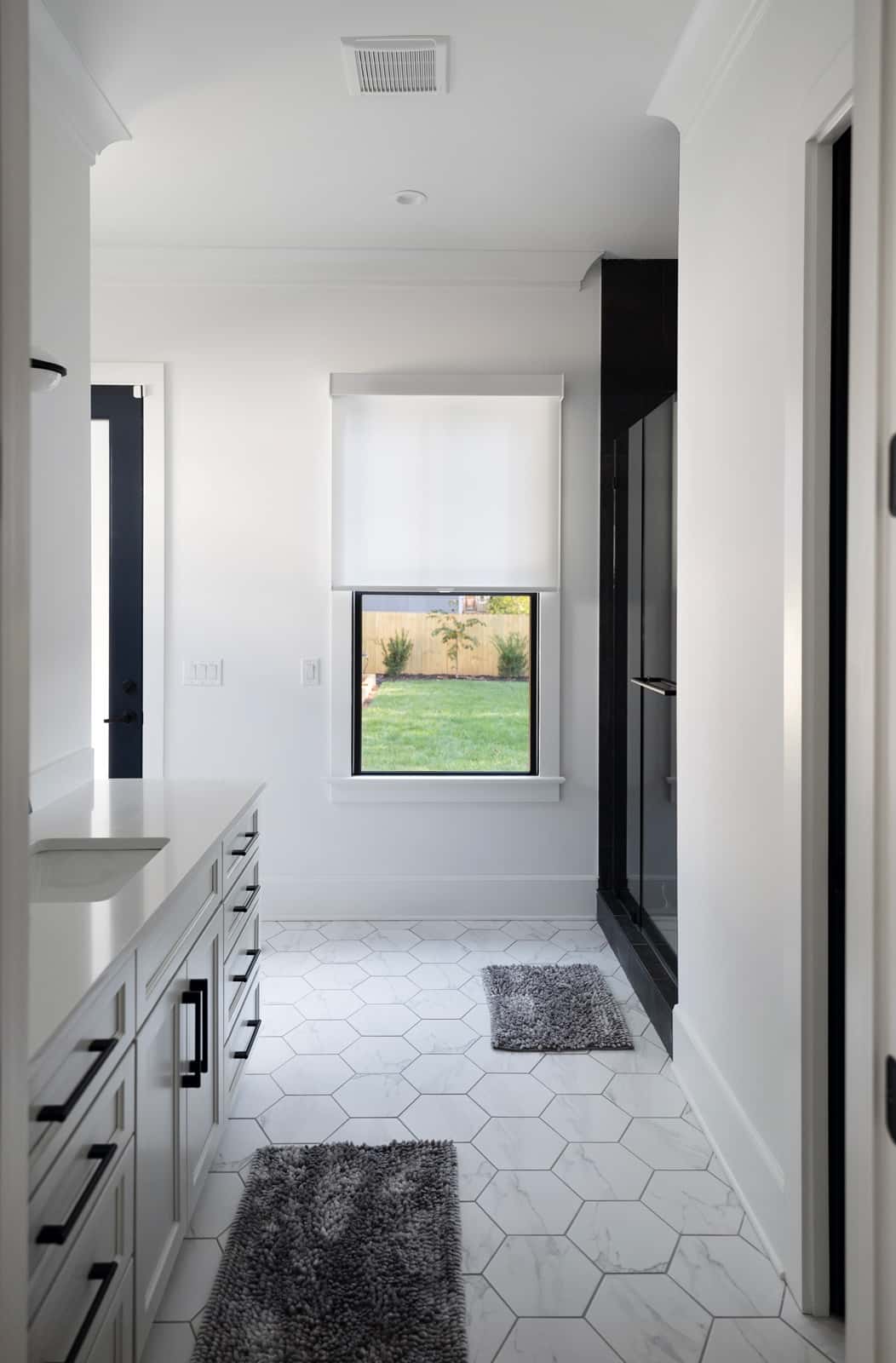Modern bathroom with white walls, a white roller window shade, hexagonal tiled floor, and a view of a backyard through the window.